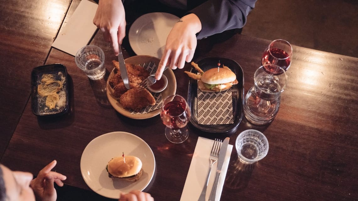 Overhead view of a wooden table set for a meal with sliders, fried chicken, glasses of red wine, and water. Hands are reaching for food, creating a cozy dining atmosphere.