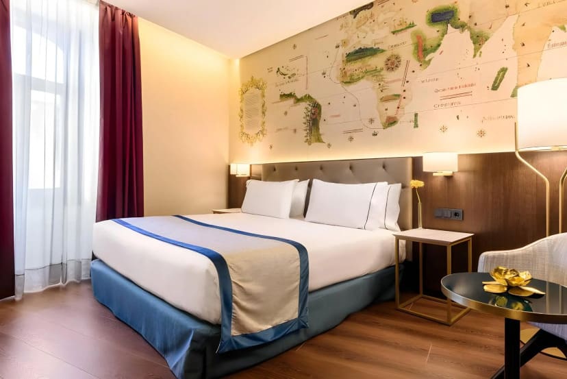 Modern hotel room with a large bed, white and blue bedding. Wall decorated with a vintage map. Soft lighting, burgundy curtains, cozy ambiance.