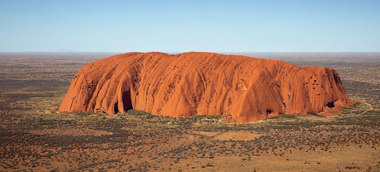Aerial view of large red plateaued rock under a blue sky
