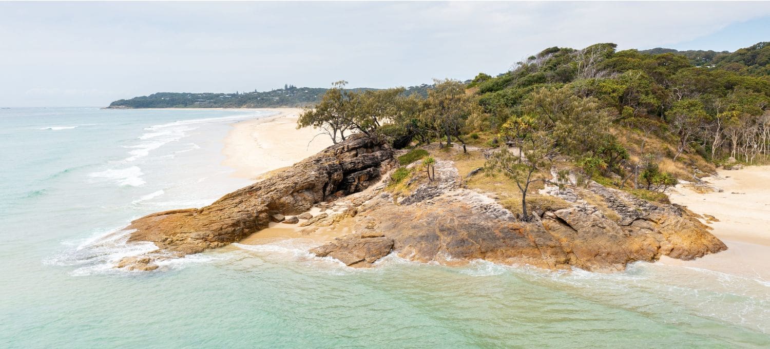 Aerial view of a serene beach, featuring a rocky outcrop covered with lush trees. Turquoise waves lap gently against the sand under an overcast sky.