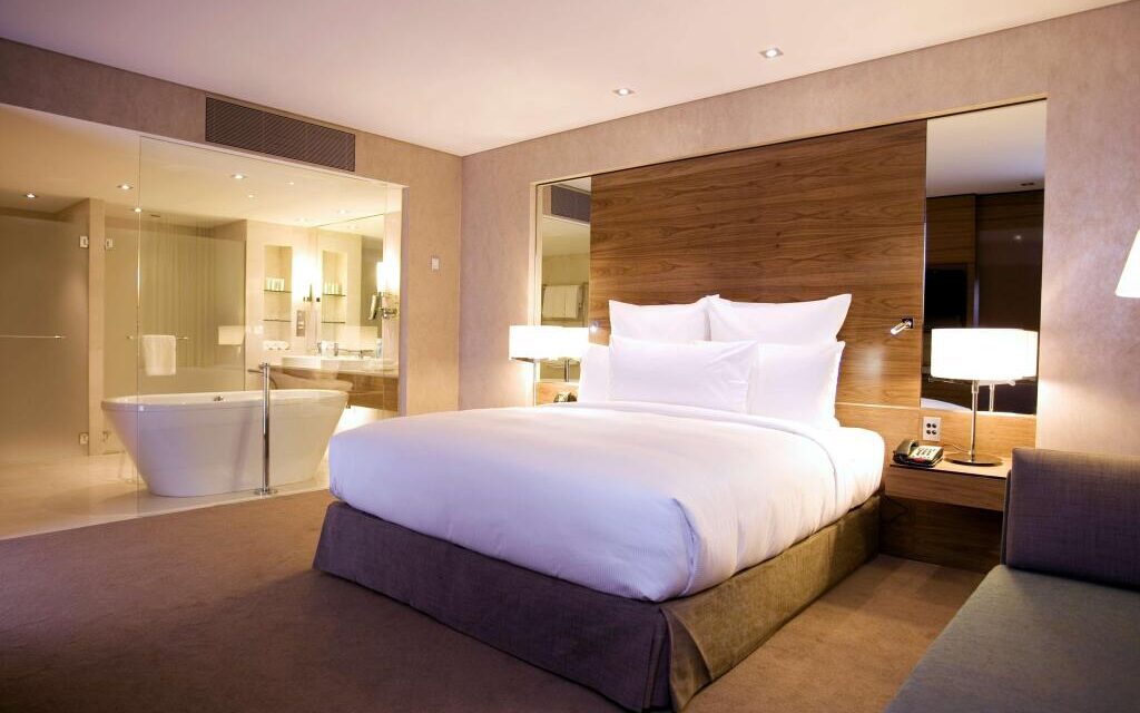 Modern hotel room with a king-sized bed and plush white bedding. Features a glass-walled bathroom with a freestanding tub, creating a luxurious feel.