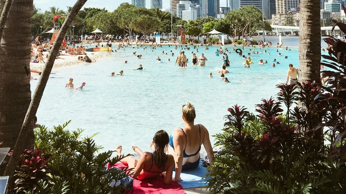 People relax by a busy urban beach lagoon on a sunny day. Two women recline on towels in the foreground, with city buildings visible in the background.