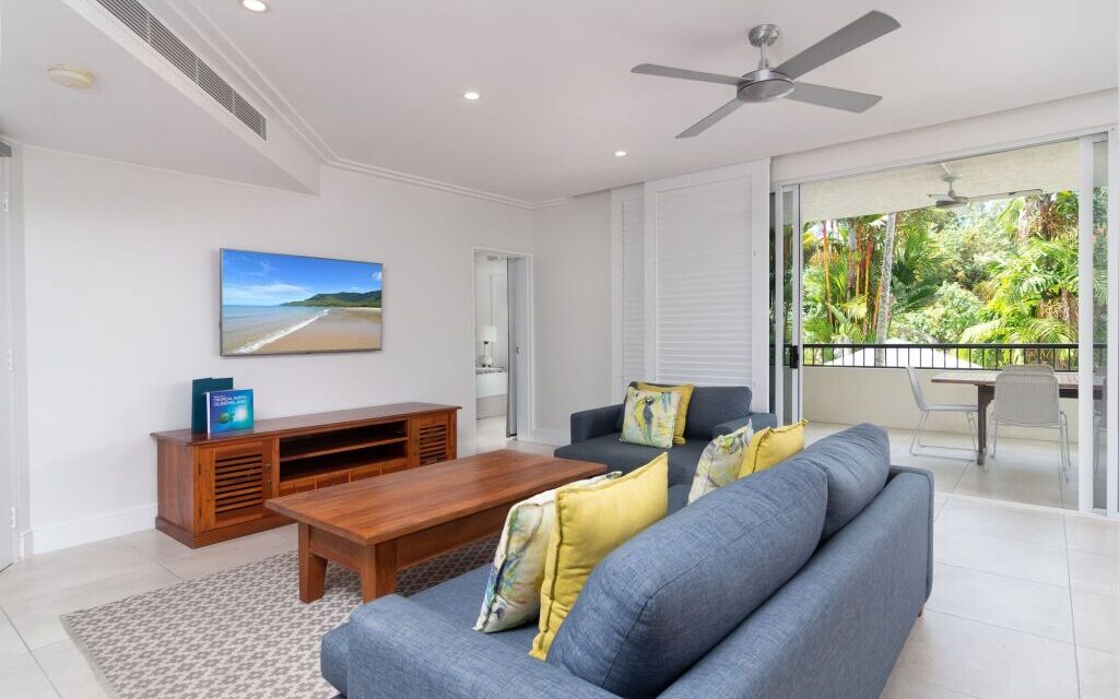 Modern living room with gray sofas and colorful pillows. A wooden coffee table faces a mounted beach scene TV. Open sliding doors lead to a balcony with lush greenery.