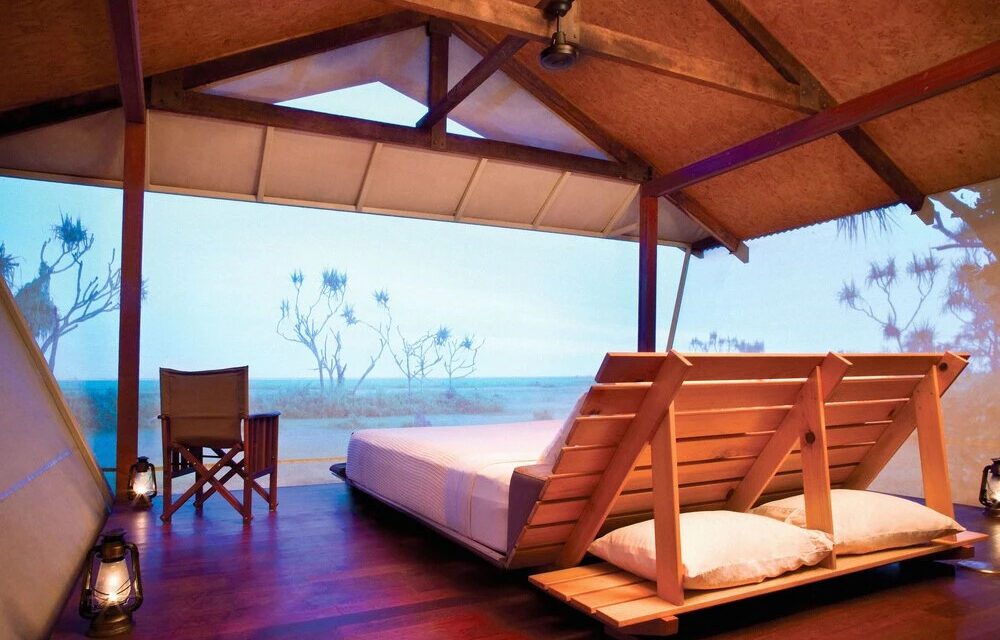 Open-air wooden bedroom facing a serene ocean view, featuring a large bed, rustic wooden chair, soft lighting from lanterns, and a calming, tranquil ambiance.