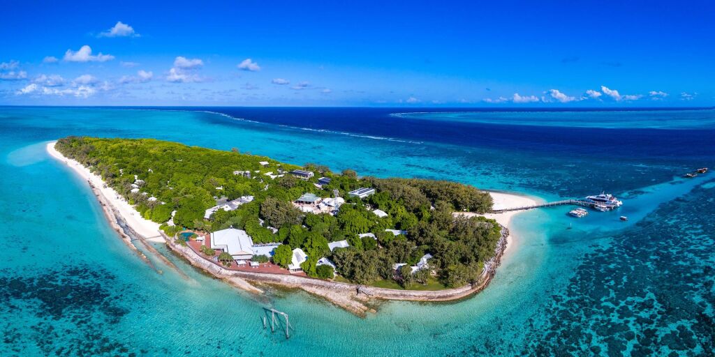 Aerial view of a lush, green tropical island surrounded by vibrant turquoise waters. White sandy beaches line the shore, and several boats are docked nearby.