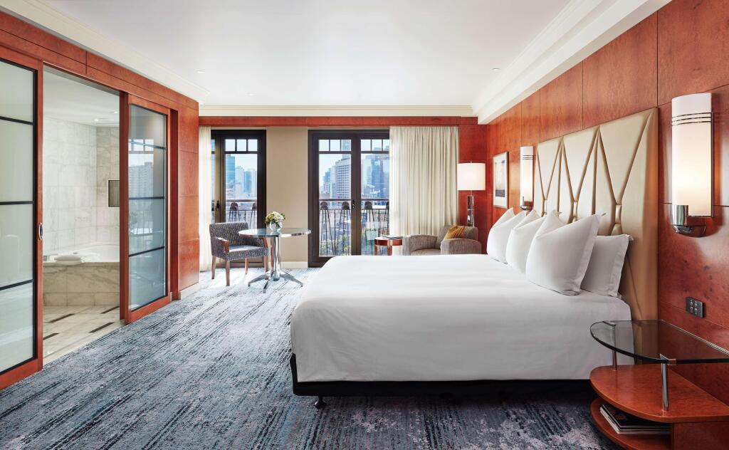 Luxurious hotel room with a large bed, elegant wood accents, and a modern design. Features a sitting area, balcony, and view of a cityscape.