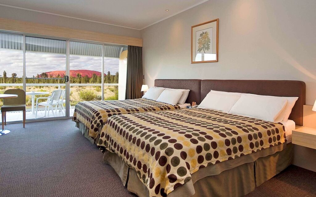 Modern hotel room with two double beds covered in polka dot blankets. A glass door opens to a patio with chairs, revealing a scenic view of a red rock formation under a cloudy sky, evoking calm and relaxation.