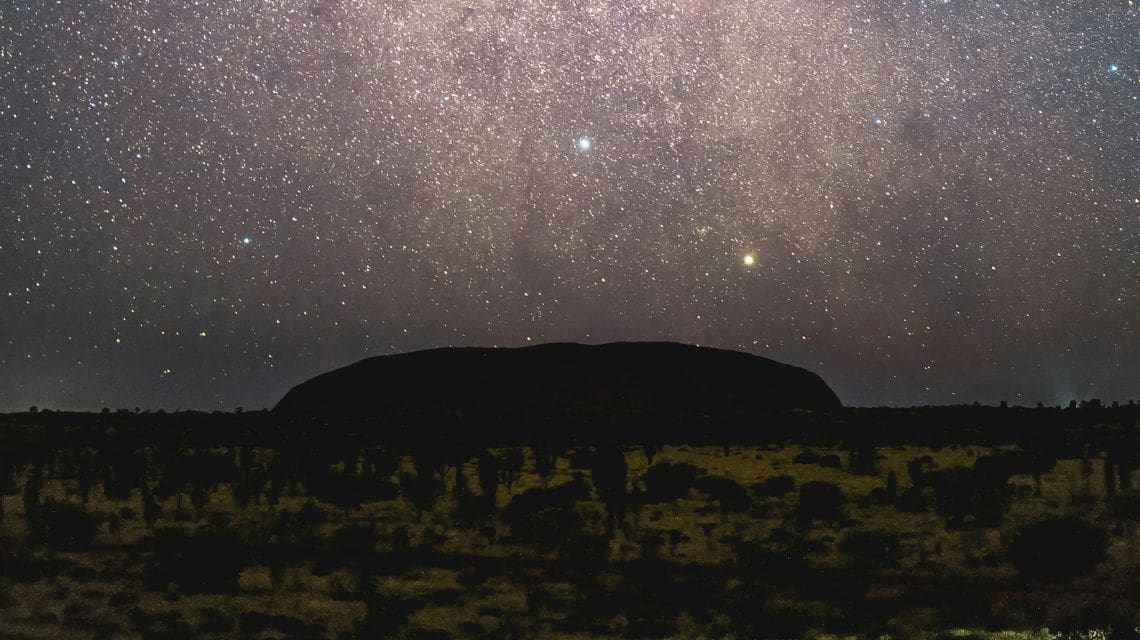 Star-filled sky over a silhouette of a large rock formation, likely Uluru, at night. The scene evokes a serene and awe-inspiring atmosphere.