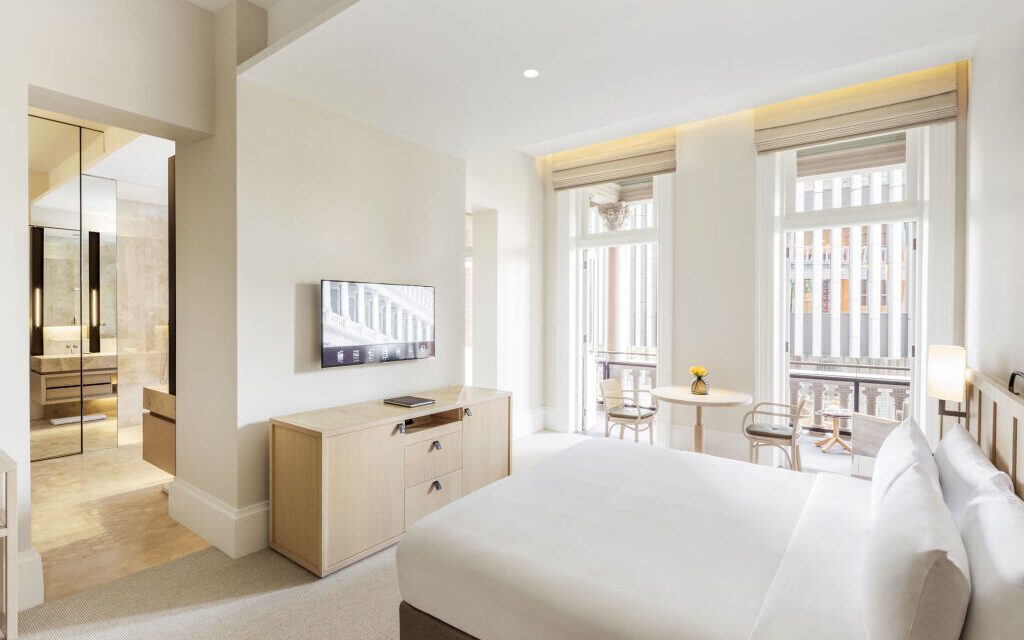 Bright hotel room with a modern, minimalist design. Features a large bed, flat-screen TV, wooden furniture, and a sitting area by tall windows. Serene and airy ambiance.