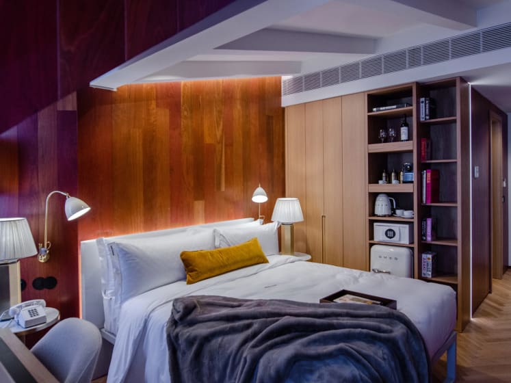 A cozy hotel room with warm wooden walls, a plush bed with white linens, and a mustard throw pillow. Soft lighting, a gray blanket, and a shelf with decor add comfort.