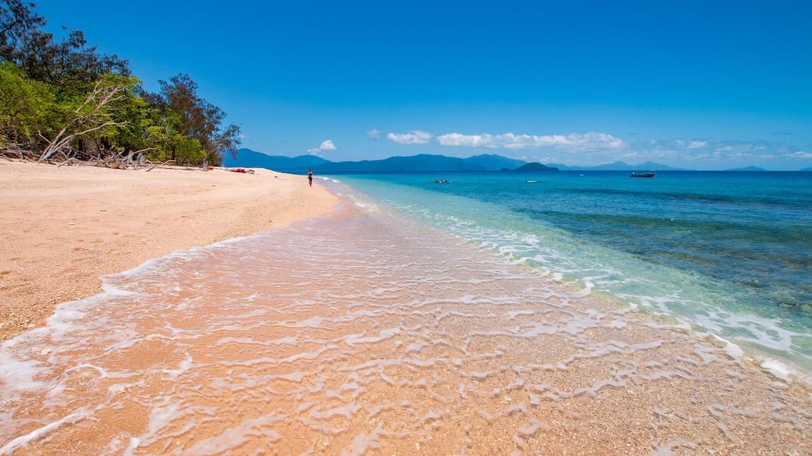 Clear ocean water and sandy beach with lush vegetation, and mountains in the background