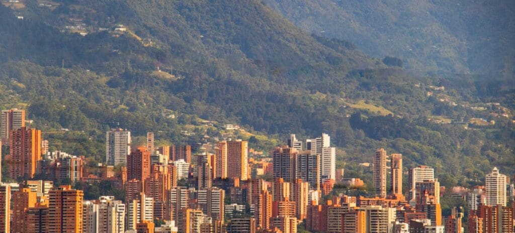 City skyline with tall, orange-hued buildings against lush, green mountains. The scene conveys a sense of urban life amidst natural beauty.