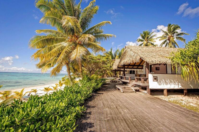Tropical beach scene with a thatched-roof hut, palm trees, and a wooden walkway beside a sandy shore. The sky is clear blue, conveying a serene, sunny atmosphere.