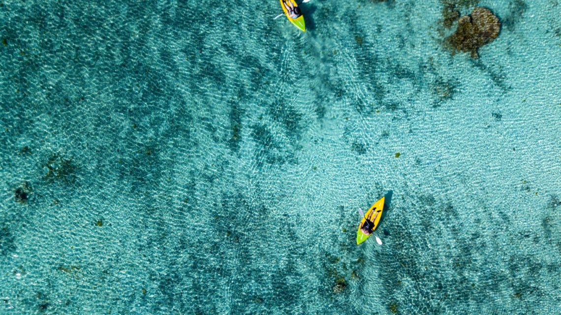 Aerial view of two people kayaking on clear turquoise water, with visible underwater rocks. The scene evokes tranquility and adventure.