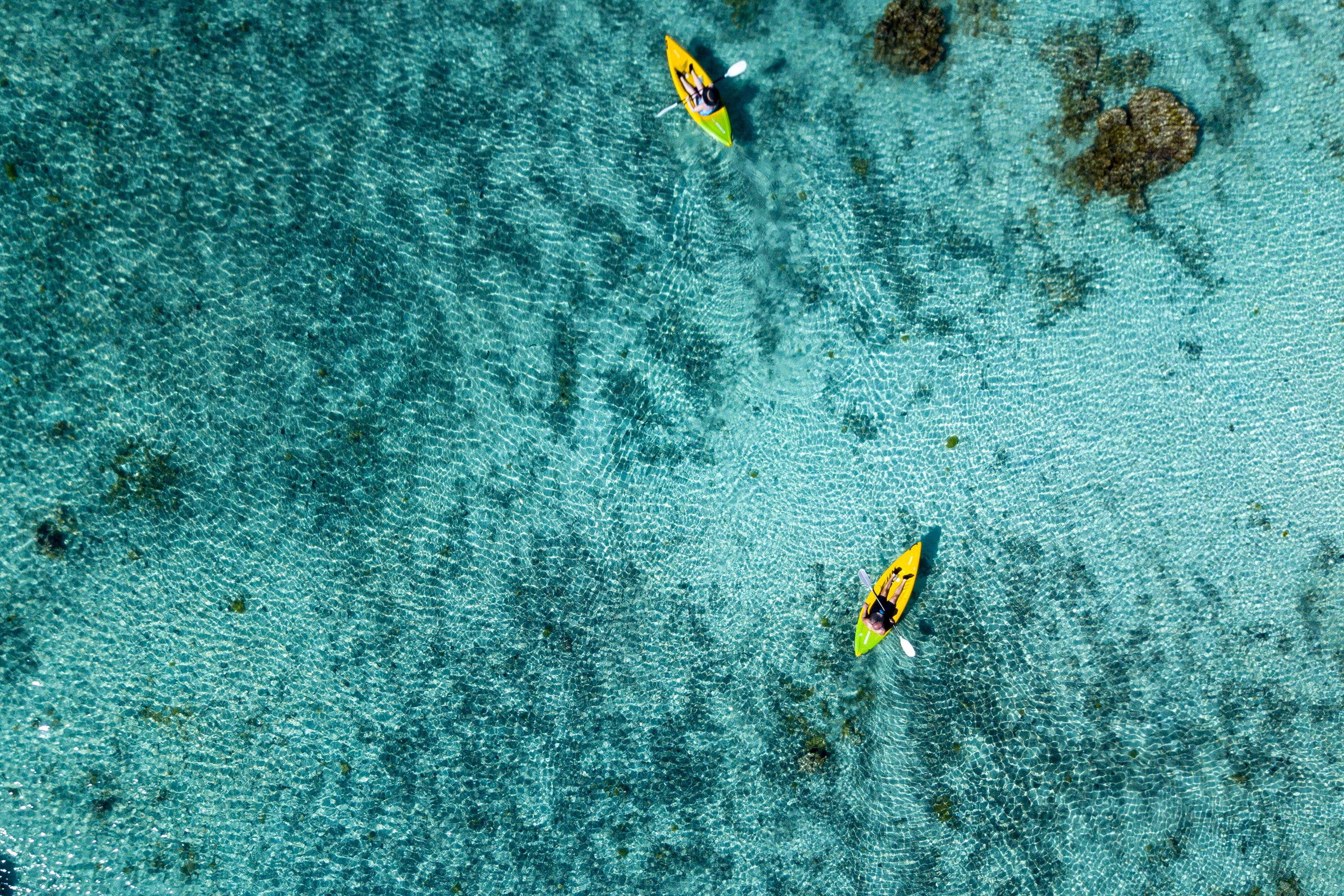 Aerial view of two people kayaking on clear turquoise water, with visible underwater rocks. The scene evokes tranquility and adventure.