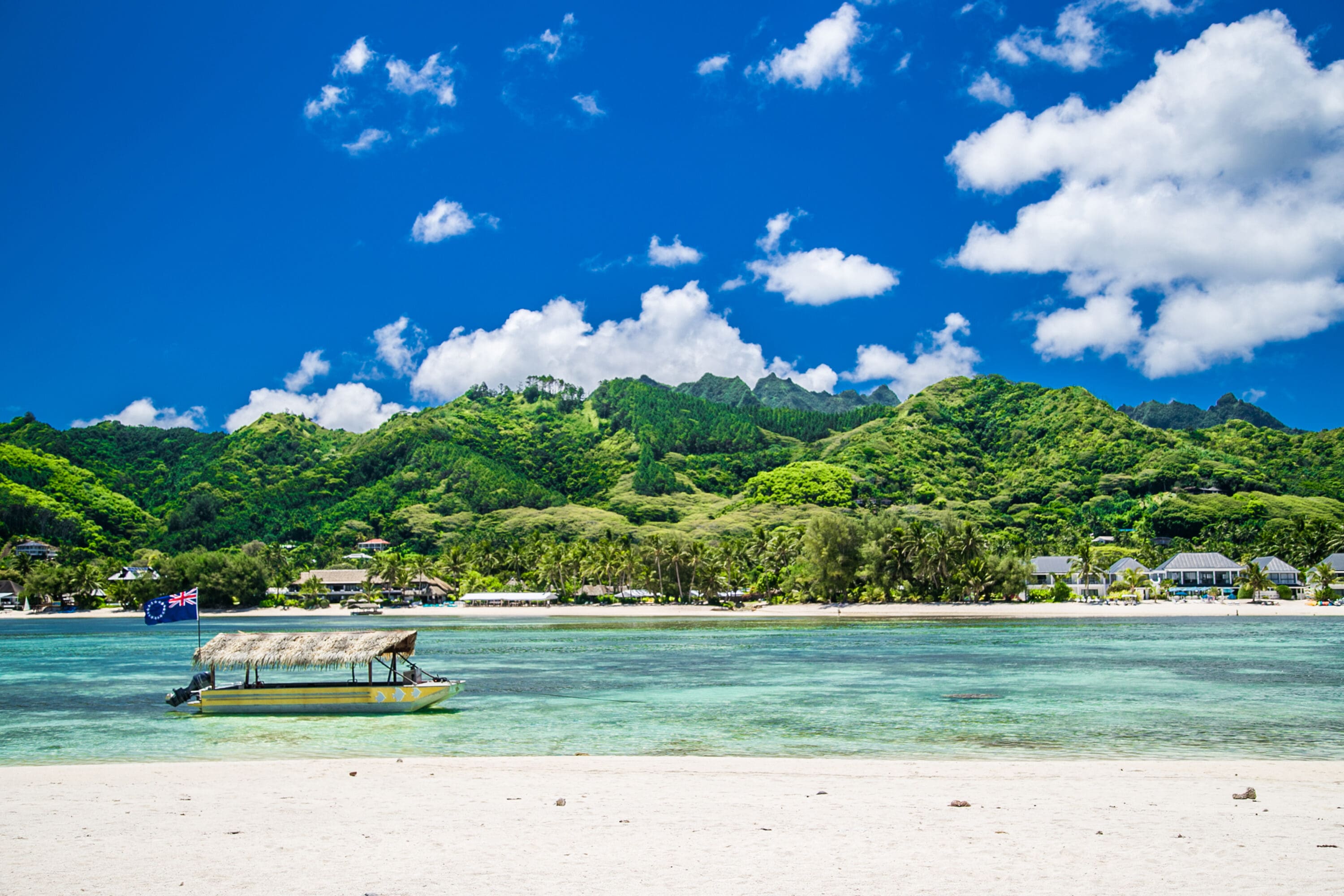 Tropical beach scene with white sand and clear turquoise water. A small thatched boat floats on the water. Lush green mountains and blue sky in the background.