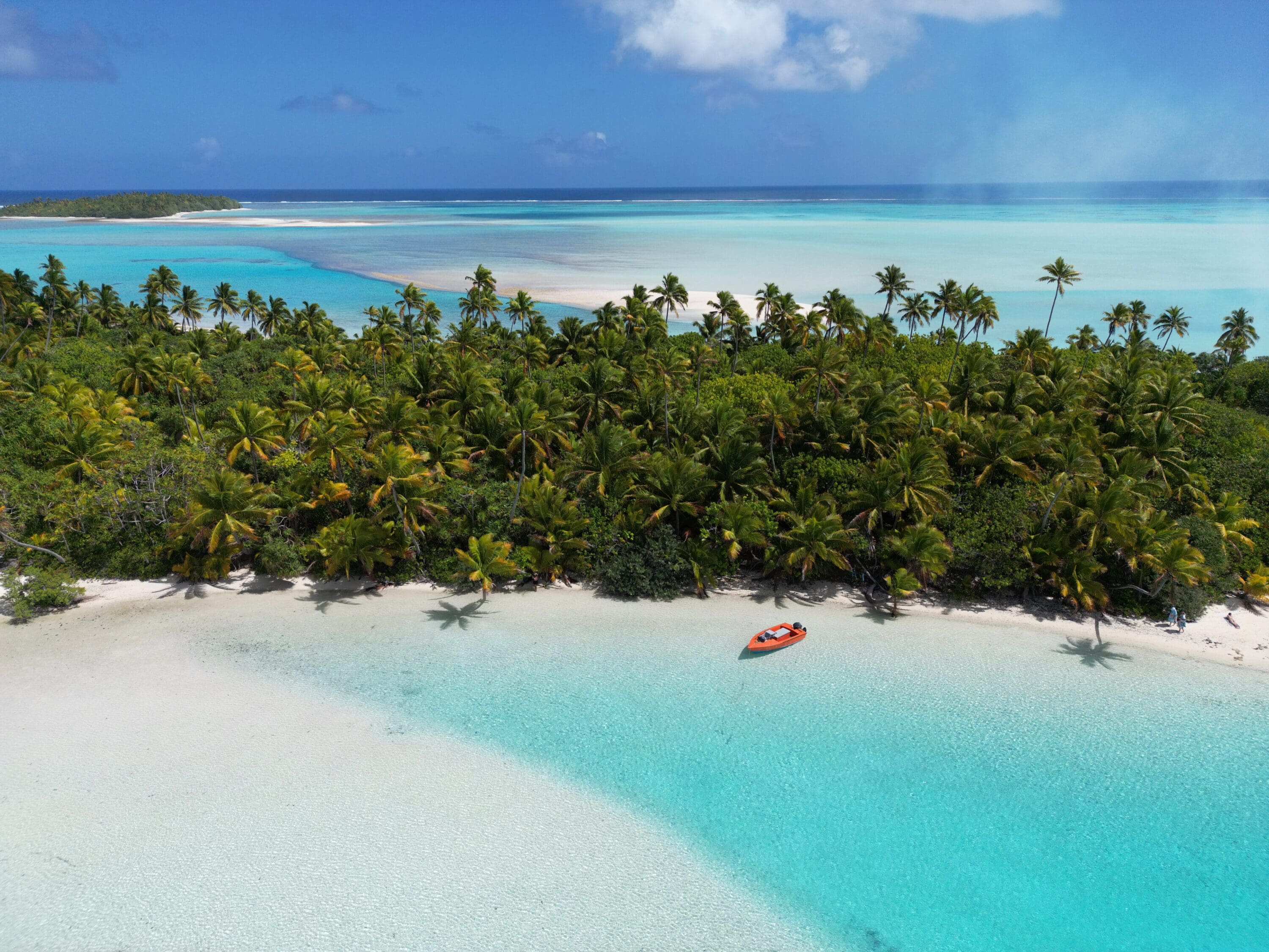 Aerial view of a tropical beach with turquoise water, white sand, and lush palm trees. A small orange boat floats near the shore under a clear blue sky.