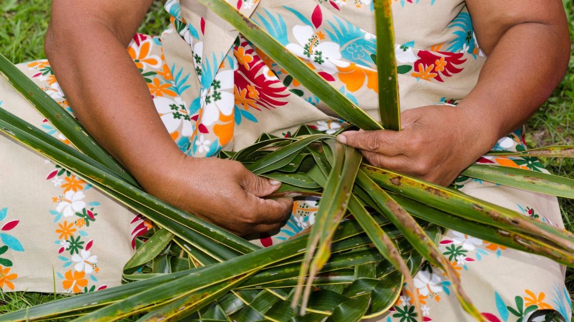 Close-up of hands weaving green palm leaves, against a backdrop of a floral dress. The mood is calm and focused, highlighting traditional craft.