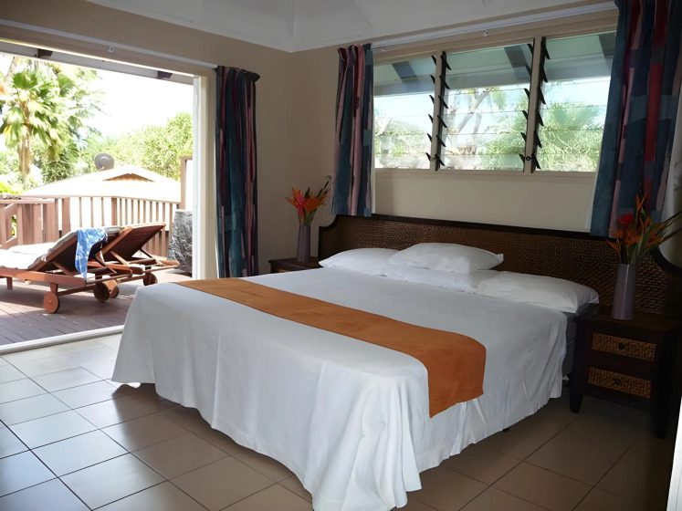 A cozy bedroom with a large bed adorned with a white cover and orange runner. Sunlit patio outside has lounge chairs, evoking a relaxing, tropical vibe.