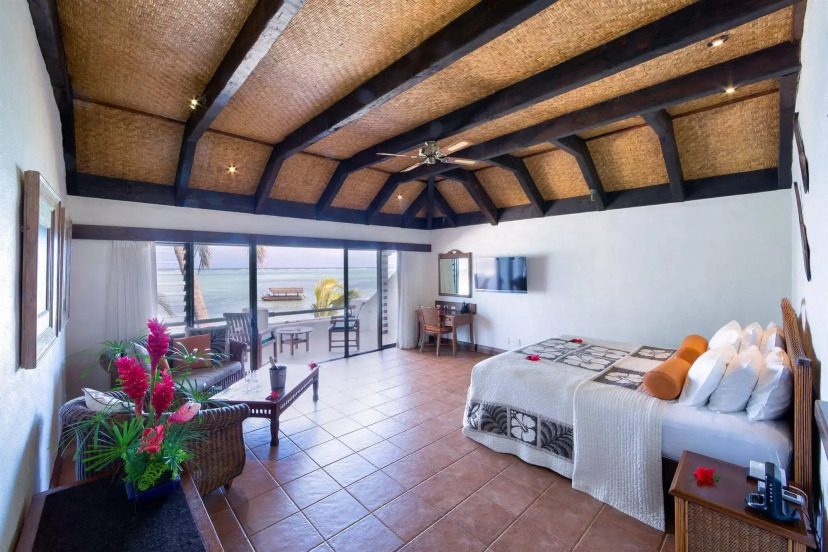 A spacious, tropical-themed bedroom with wooden ceiling beams, a large bed with decorative pillows, and a patio with ocean views. Bright and inviting ambiance.