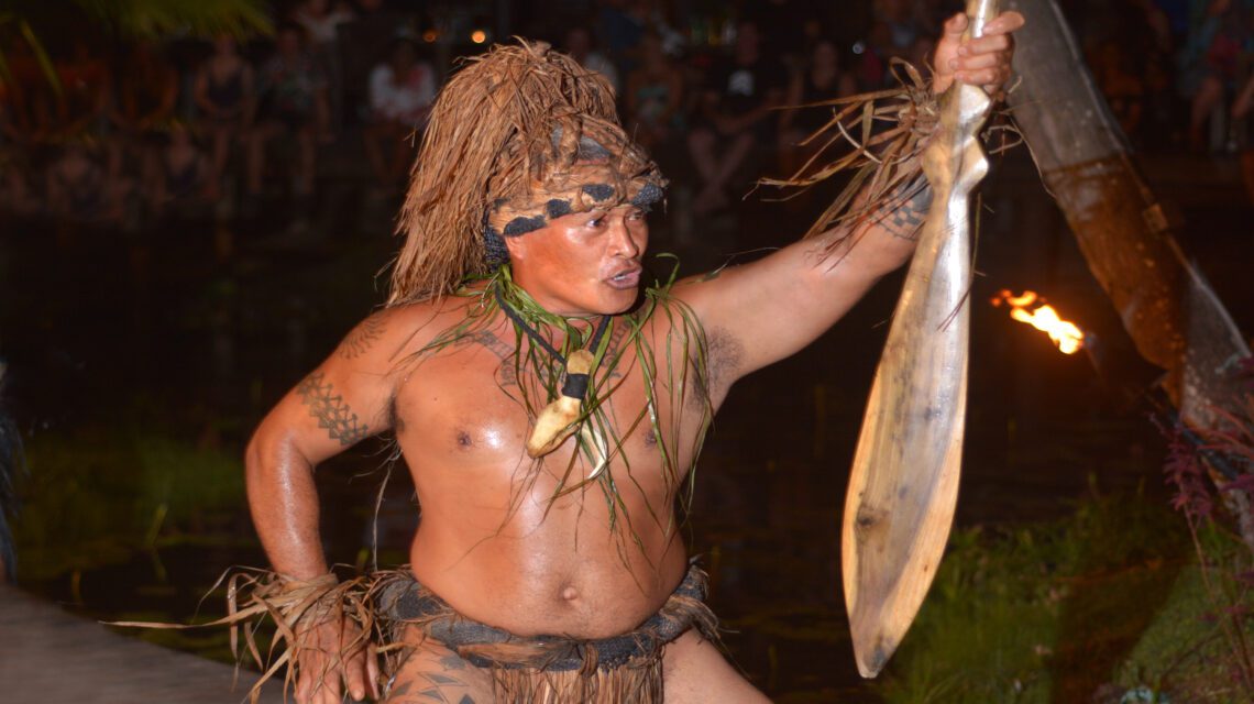 A performer in traditional attire energetically poses with a wooden paddle. He wears a headdress and grass skirt, conveying intensity and cultural richness.