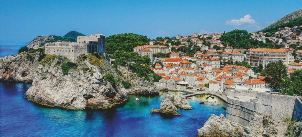 Coastal cityscape of Dubrovnik, Croatia. Features medieval stone fortresses atop rocky cliffs, orange-tiled rooftops, and vibrant blue sea under clear skies.