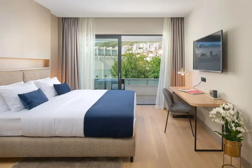 Modern hotel room with a large bed featuring blue accents, a sleek desk beside it, and a wall-mounted TV. A glass door reveals a balcony view.
