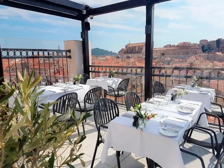 Rooftop restaurant with neatly set tables and white linens overlooking historic city rooftops under a clear blue sky, conveying a serene, elegant atmosphere.