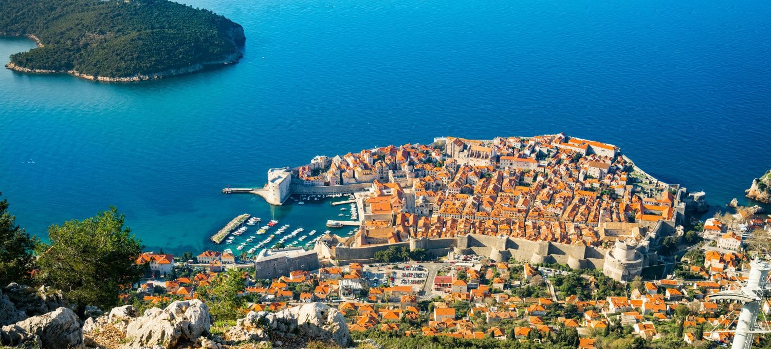 Aerial view of Dubrovnik, Croatia, showing the historic old town with its iconic orange-tiled roofs surrounded by medieval walls, next to the blue Adriatic Sea.