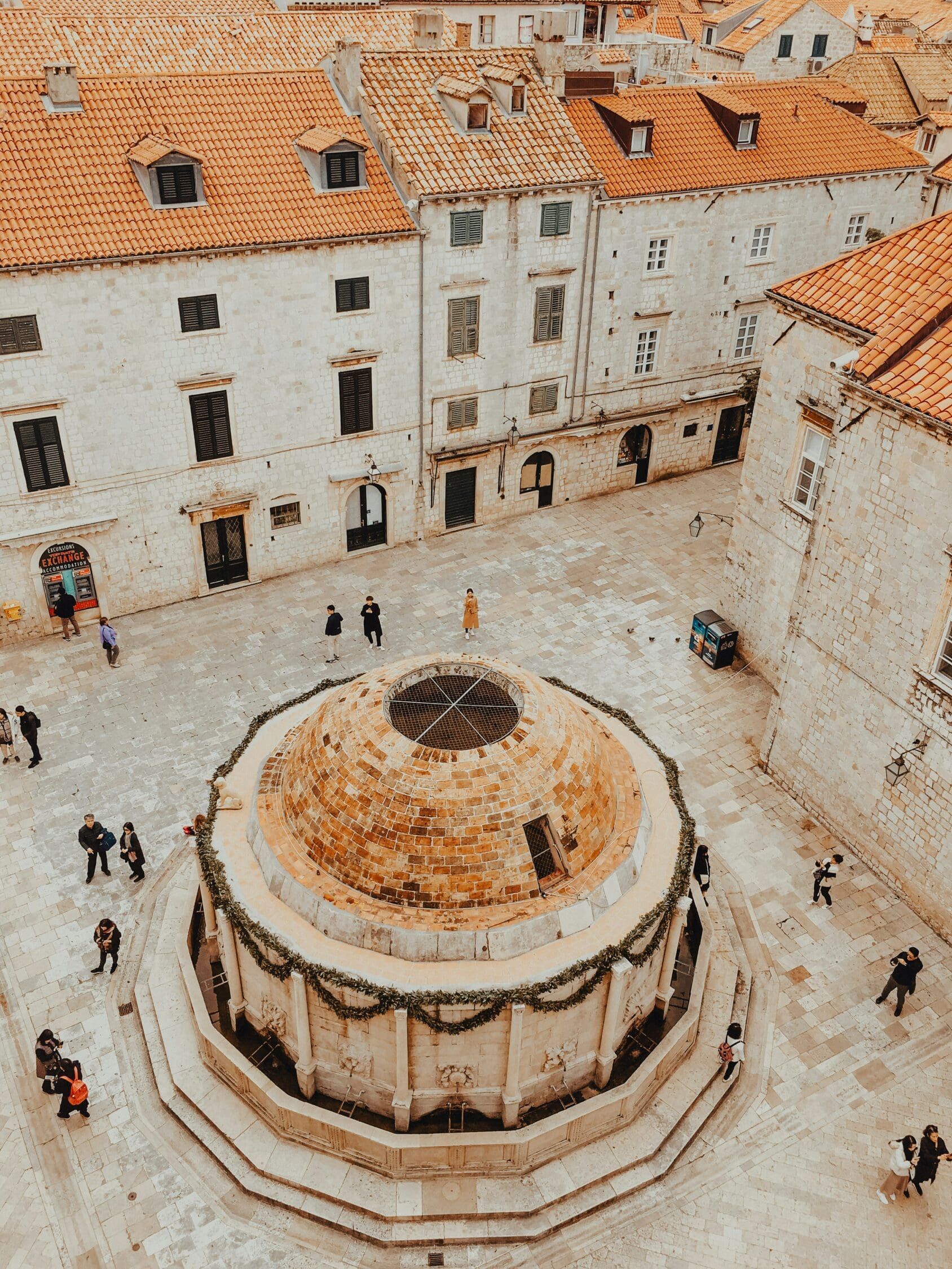 Aerial view of an old stone fountain in a medieval square, surrounded by historic buildings with red-tiled roofs. People stroll around, creating a lively atmosphere.