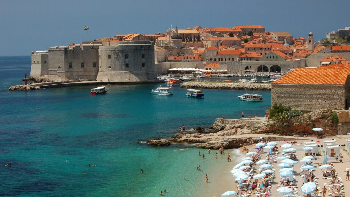 Sunlit coastal scene of Dubrovnik, Croatia, with turquoise waters, historic stone buildings, and orange rooftops. Crowded beach with umbrellas. Serene atmosphere.
