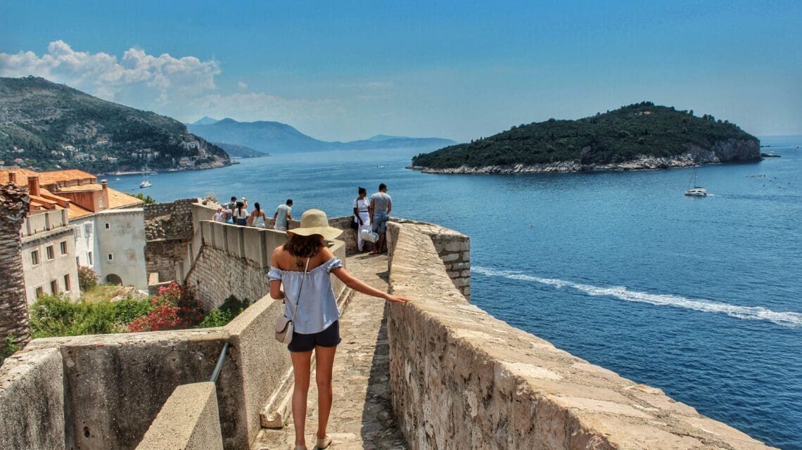 A woman in a hat walks along a stone wall overlooking the sea, with an island in the distance. Clear blue skies suggest a peaceful, sunny day.