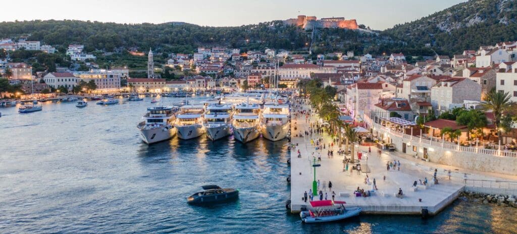 A scenic harbor at dusk with yachts docked along a vibrant promenade bustling with people. Historic buildings and a lit fortress overlook the water.
