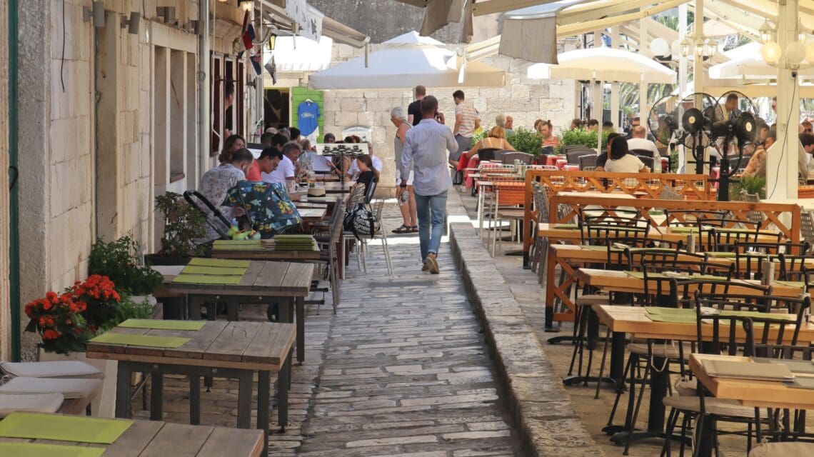 A narrow cobblestone street with outdoor cafes on both sides, tables set with green placemats. People dining, a relaxed and inviting atmosphere under large awnings.