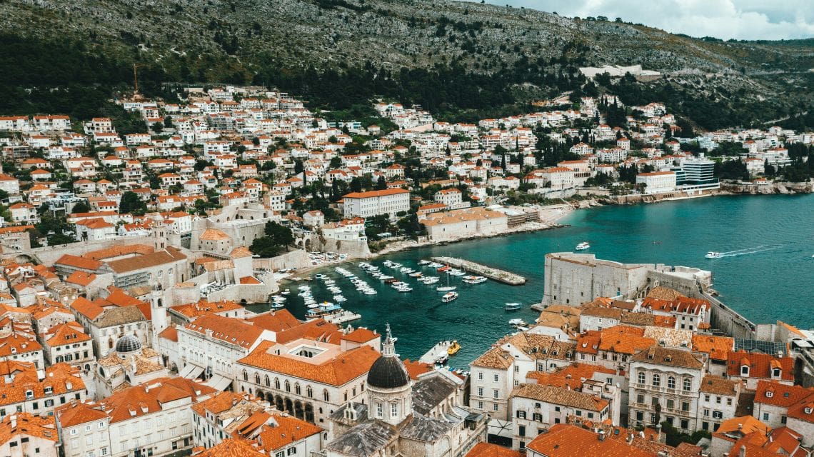 Aerial view of Dubrovnik, Croatia, showcasing red-roofed buildings, a historic fortress, turquoise sea, and boats, set against a green hillside backdrop.