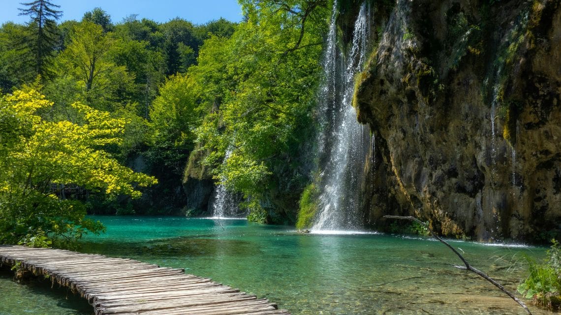 A serene waterfall cascades into a turquoise lake surrounded by lush greenery and tall trees. A rustic wooden path leads across the water. Tranquil and natural.