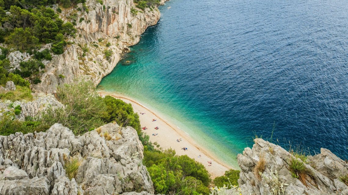 A secluded beach with golden sand nestled between rocky cliffs and lush greenery. The turquoise water meets deep blue, creating a serene coastal scene.