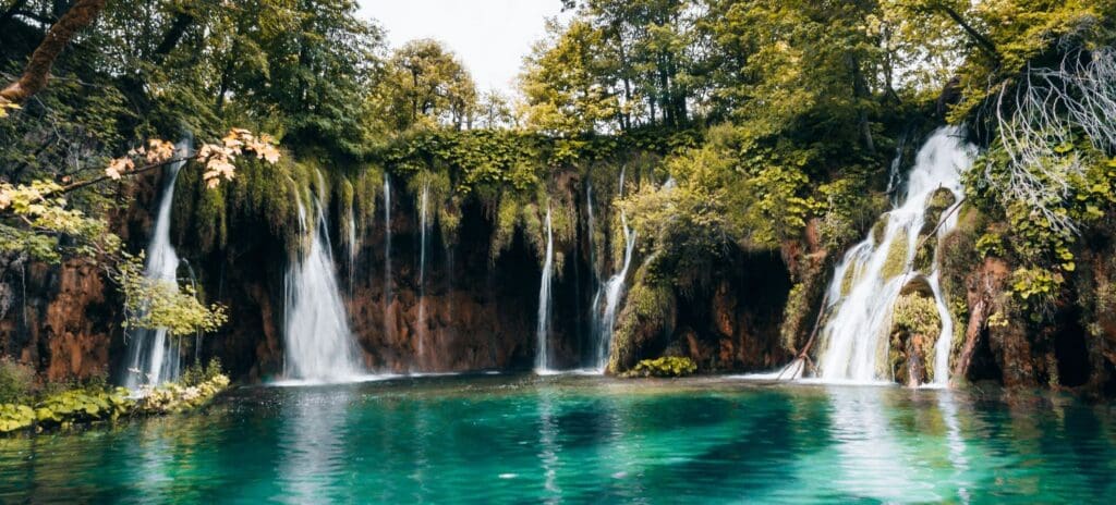 A serene scene of cascading waterfalls flowing over lush, green cliffs into a turquoise pool, surrounded by dense trees, conveying tranquility and natural beauty.