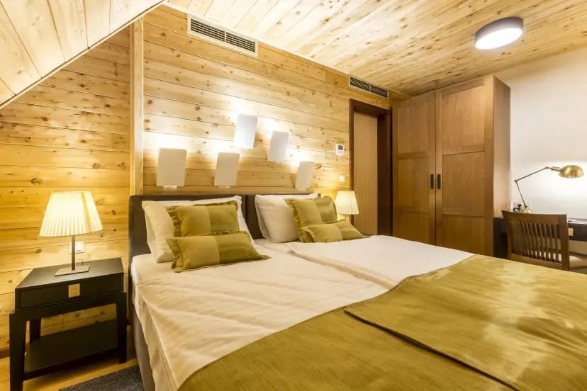A cozy bedroom with wooden walls and ceiling features two neatly made twin beds with green accents. Soft lighting and a desk create a warm, inviting ambiance.