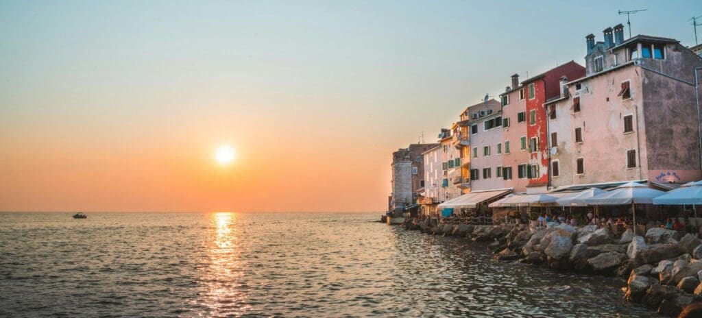 Coastal town at sunset, with colorful buildings lining a rocky shore and outdoor cafes. The sun sets over calm sea, creating a peaceful, warm atmosphere.
