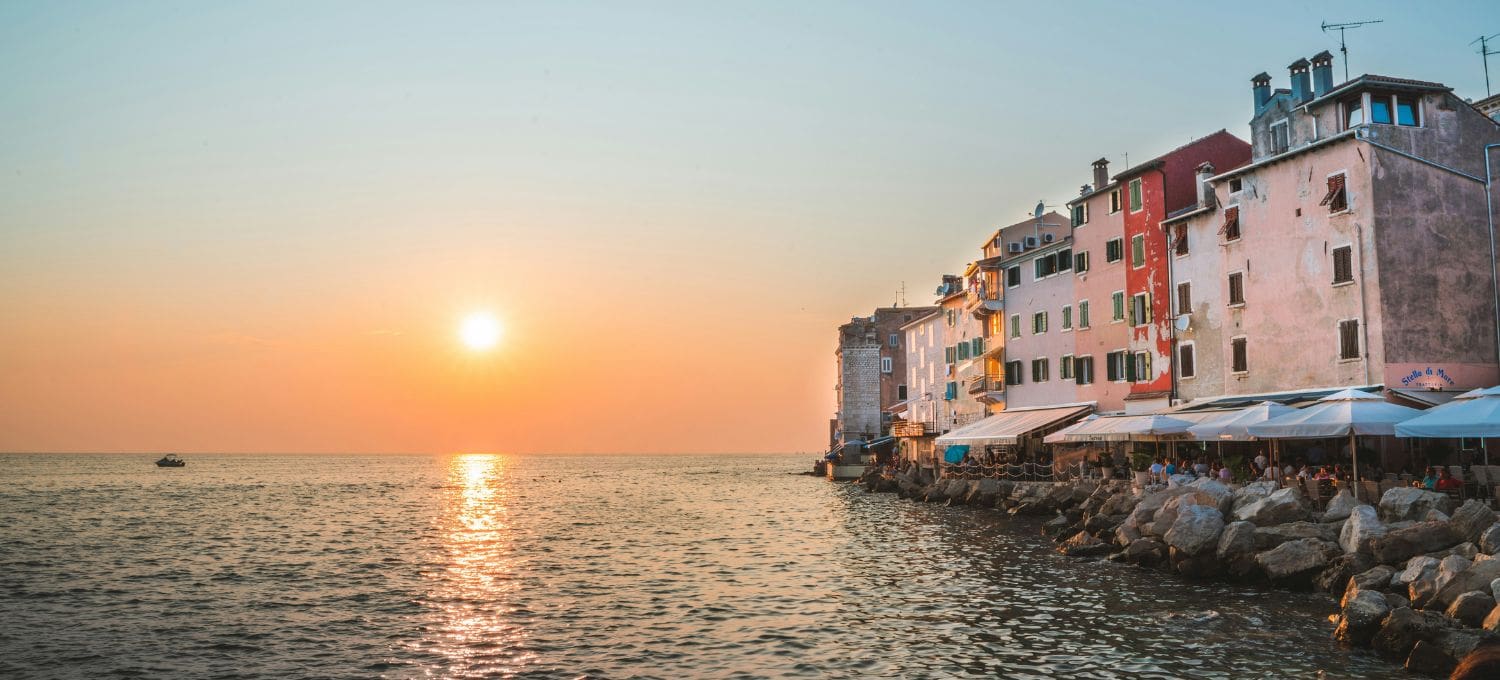 Coastal town at sunset, with colorful buildings lining a rocky shore and outdoor cafes. The sun sets over calm sea, creating a peaceful, warm atmosphere.