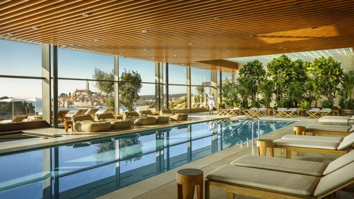Luxurious indoor pool with sun loungers, potted plants, and wooden ceiling. Large windows reveal a seaside view and distant town, creating a serene ambiance.
