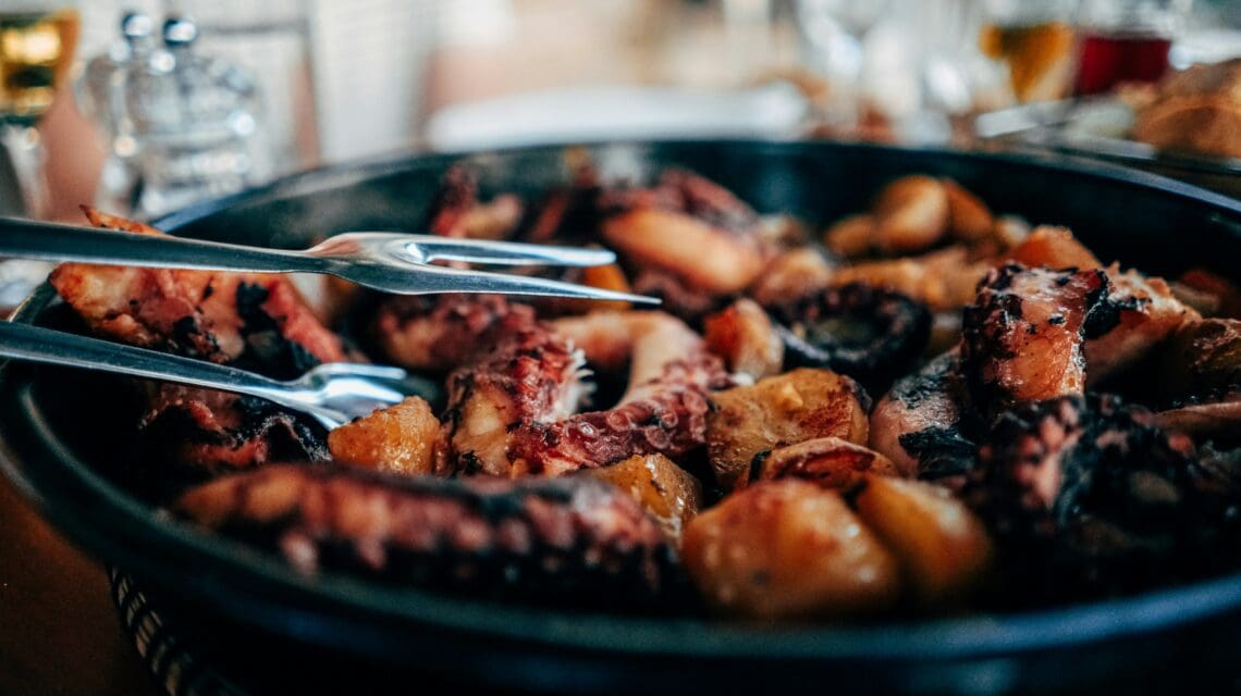 A close-up of a dish featuring grilled octopus and potatoes served in a pan. The focus on the food gives a warm, inviting atmosphere.
