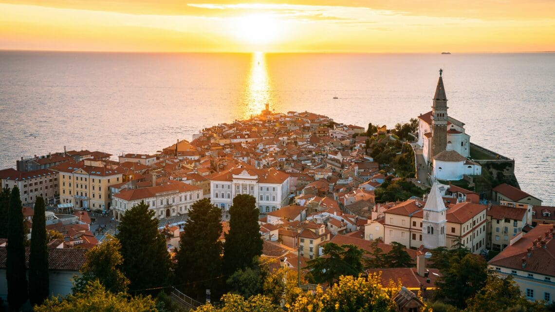 Sunset over Piran, Slovenia. The golden sun reflects on the sea, illuminating red-roofed coastal buildings and a prominent church tower, evoking tranquility.