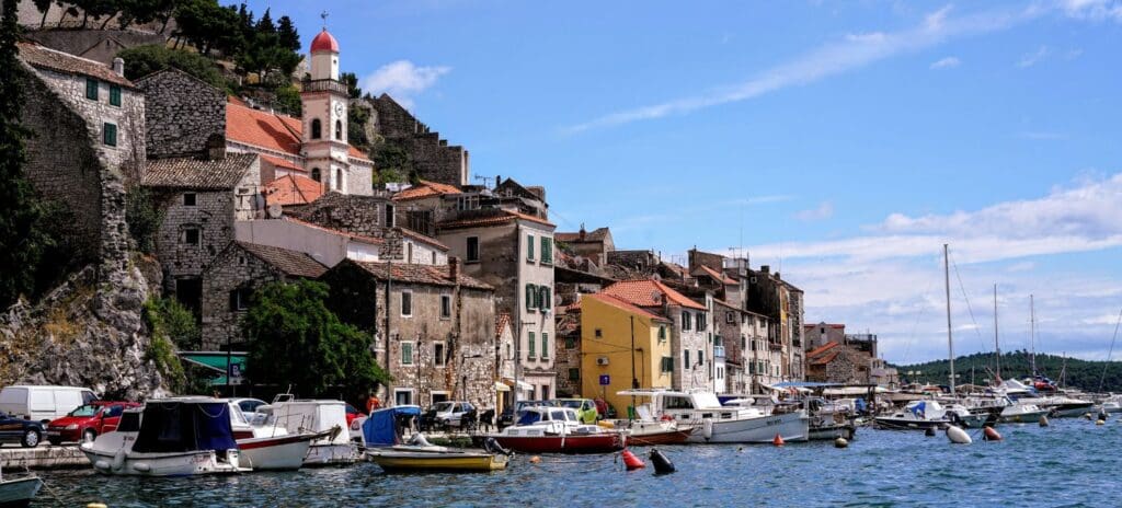 Scenic coastal town with rustic stone buildings and a white-domed church. Boats dock along the blue water, and a vibrant sky enhances the serene atmosphere.
