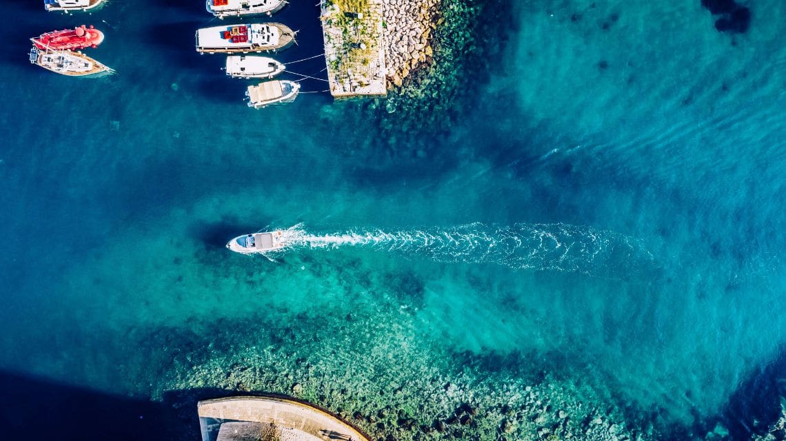 Aerial view of a small boat moving through clear blue water, leaving a trail. Docked boats and a stone pier with greenery are visible at the top, with a calm, serene atmosphere.