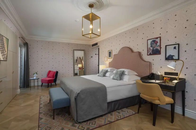 A cozy hotel room with a large bed, gray and pink decor, a modern chandelier, a desk with a yellow chair, a pink armchair, and framed art on the wall.