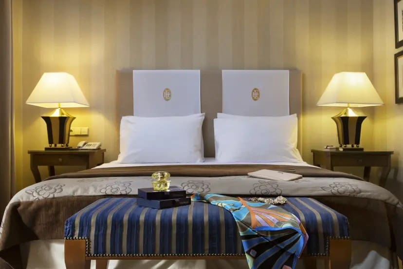 Elegant hotel room with a king-sized bed, crisp white pillows, and gold bedside lamps on wooden tables. A luxurious bed scarf and decorative items adorn the bed foot.