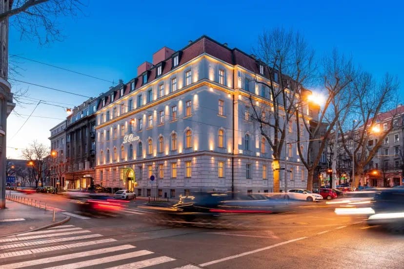 Street corner with a grand, illuminated building at dusk. Blurred car lights suggest motion, while bare trees and a deep blue sky create a calm urban scene.