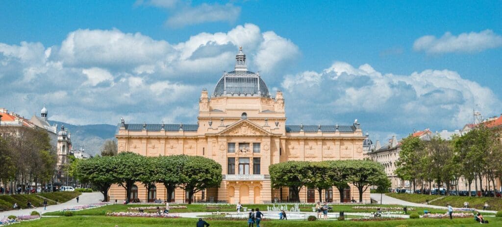 Historic yellow building with a domed roof surrounded by manicured gardens and trees under a blue sky with scattered clouds; visitors walk in the park.